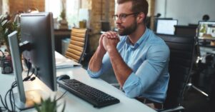 Thinking. Side view of young bearded man in eyeglasses and formal wear working on computer and keeping palms pressed together while sitting in the modern office