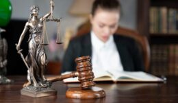 Young female lawyer during work in chamber. Gavel and Themis statue on the brown shining desk.